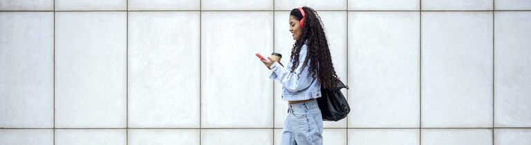 Foto einer jungen Frau in Jeans-Klamotten, die auf ihre Handy schauend mit einem Kaffe-to-Go-Becher in der Hand die Straße entlang läuft