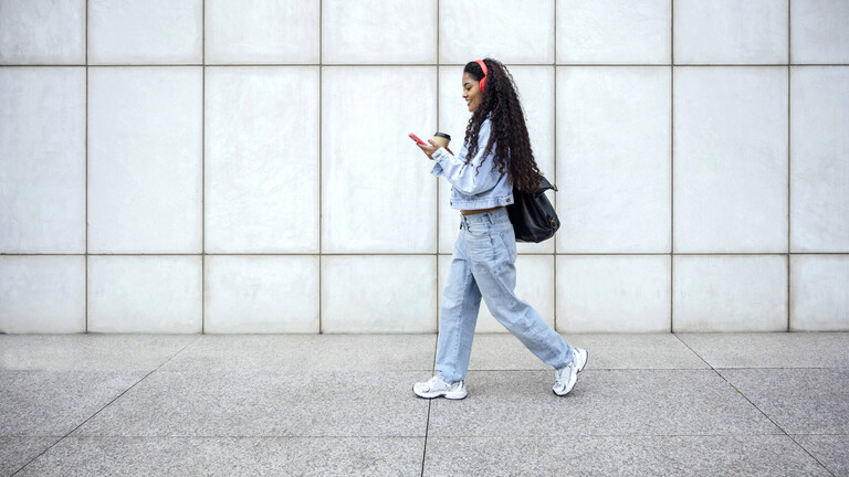 Foto einer jungen Frau in Jeans-Klamotten, die auf ihre Handy schauend mit einem Kaffe-to-Go-Becher in der Hand die Straße entlang läuft