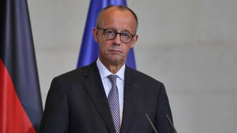 Bundeskanzler Friedrich Merz (CDU) steht vor Mikrofonen und blickt ernst in die Kamera. Im Hintergrund ist deutsche und die Flagge der EU zu sehen.