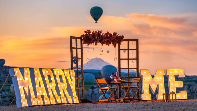 Große leuchtende Schriftzeichen "Marry Me" stehen am Strand, inklusive Holrahmen und dekoriertem Tisch mit Stühlen