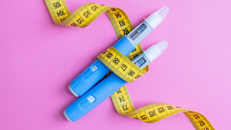 Zwei blaue Insulinstifte, umwickelt mit einem gelben Maßband auf rosa Hintergrund, symbolisieren Diabetes-Management, Gewichtsabnahme, medizinische Behandlung und Gesundheitskonzepte.