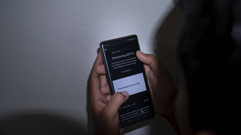 Person hält ein Smartphone und liest über GPT-5 vor einer weißen Wand bei schwachem Licht.