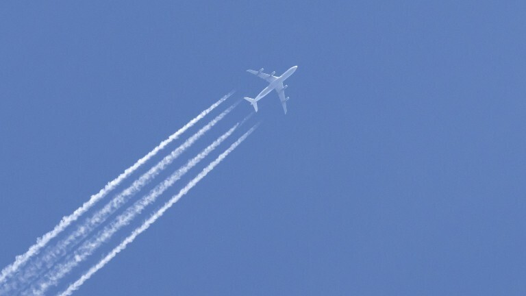 Am blauen, wolkenlosen Himmel ist ein Flugzeug zu sehen, das Kondensstreifen hinterlässt.