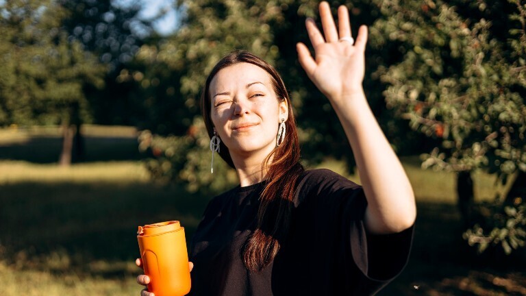 Eine Frau ist an einem sonnigen Tag draußen unterwegs. Sie hält einen orangefarbenen Thermobecher in der einen Hand und die andere Hand vor ihr Gesicht, um sich vor der Sonne zu schützen.