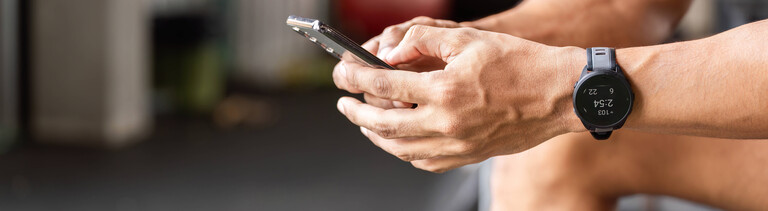 Ein Sportler nutzt eine Fitness-App auf seinem Smartphone während einer Pause im Fitnessstudio. Er sitzt dort, konzentriert auf sein Smartphone und verfolgt mithilfe einer Fitness-App seine Fortschritte und Trainingseinheiten.