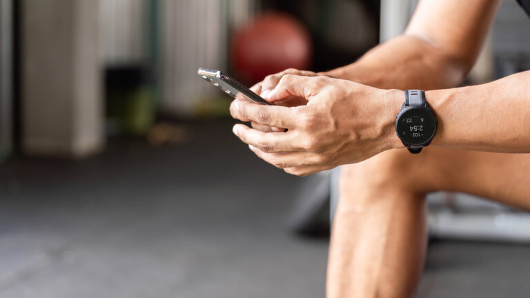 Ein Sportler nutzt eine Fitness-App auf seinem Smartphone während einer Pause im Fitnessstudio. Er sitzt dort, konzentriert auf sein Smartphone und verfolgt mithilfe einer Fitness-App seine Fortschritte und Trainingseinheiten.