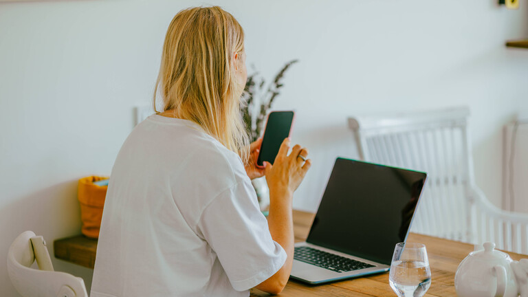 Frau an einem Schreibtisch, mit einem Smartphone in der Hand. Vor ihr auf dem Tisch ein Laptop.