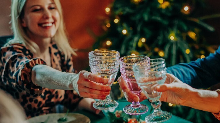 Drei Personen stoßen gemeinsam an. Sie sitzen an einem festlich geschmückten Tisch. Im Hintergrund steht ein Weihnachtsbaum.