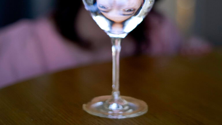 EIne Frau, deren Gesicht von einem Wasserglas verdeckt ist. Das gesicht spiegelt sich jedoch im Glas.