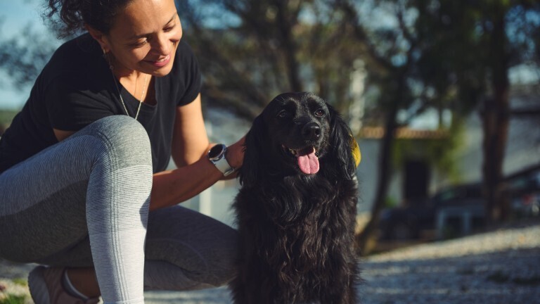 Eine junge Frau ist draußen spazieren und in einer Hocke. Sie streichelt einen Hund, der neben ihr steht.