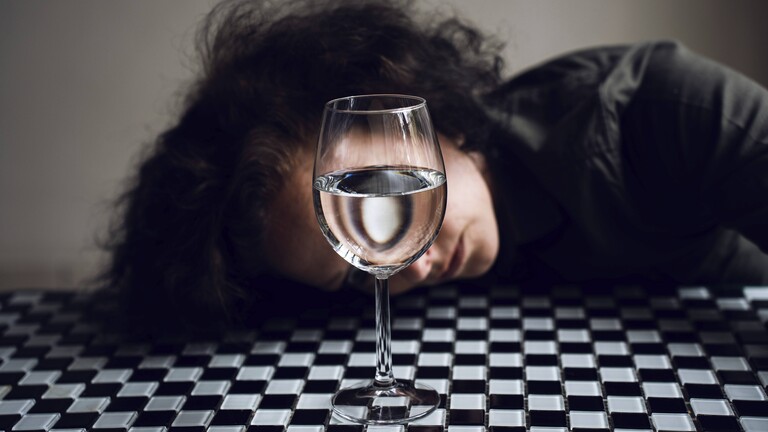 Eine junge Frau hat den Kopf auf einen schachbrettgemusterten Tisch gelegt, ihr Gesicht ist von einem mit Wasser gefüllten Weinglas verdeckt.