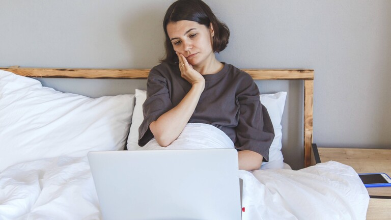 Eine junge Frau sitzt im Bett. Sie hat einen Laptop auf dem Schoß und hält sich eine Hand an ihre Wange. Sie wirkt müde und nachdenklich.