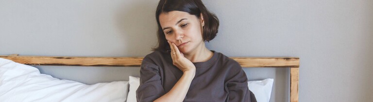 Eine junge Frau sitzt im Bett. Sie hat einen Laptop auf dem Schoß und hält sich eine Hand an ihre Wange. Sie wirkt müde und nachdenklich.