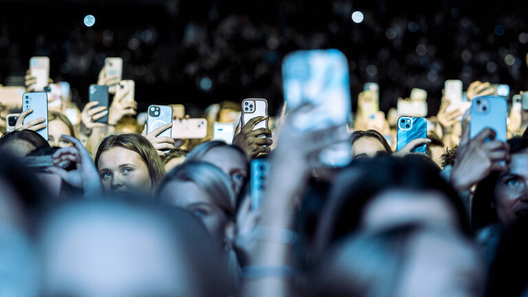 Eine Konzerthalle ist gefüllt mit Menschen. Das Publikum blickt in Richtung Bühne. Viele halten dabei ein Handy hoch.