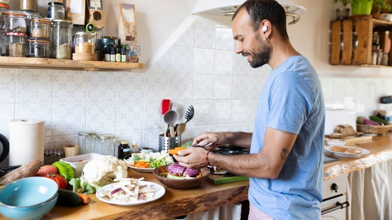 Ein Mann steht lächelnd in einer Küche und bereitet dabei Gemüse zu. Er hat einen kabellosen Kopfhörer im Ohr.