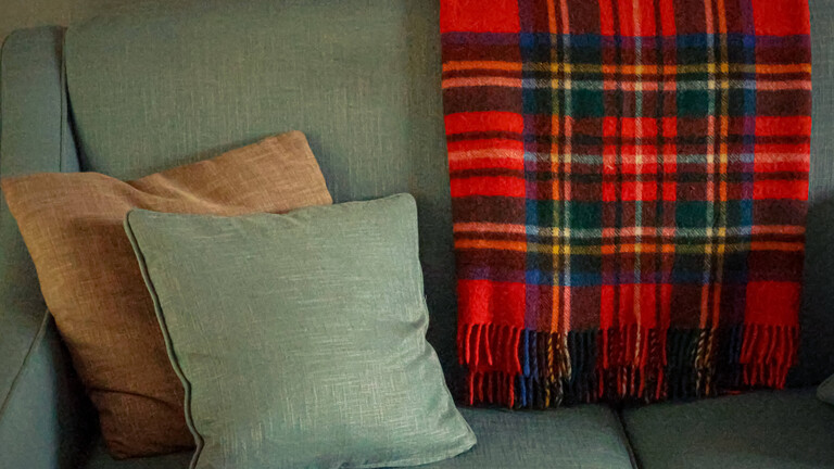 Ein Sofa mit grünem Stoffbezug und mit einem braunen und grünen Sofakissen, über der Rückenlehne liegt eine rot-schwarz karierte Wolldecke.