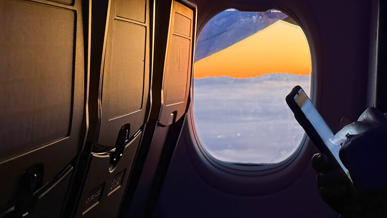 Smartphone im Flugzeug mit Blick auf Abendstimmung aus dem Flugzeugfenster