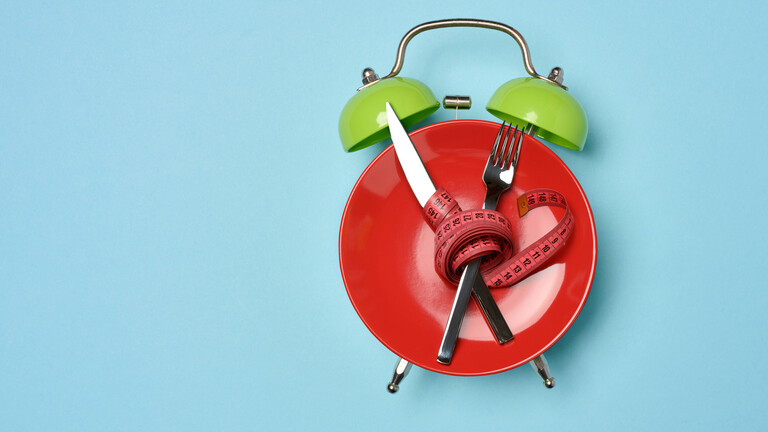 Diät-Konzeptbild, roter Teller, der zum Wecker stilisiert wurde, mit gekreuztem Besteck umwickelt mit rotem Maßband
