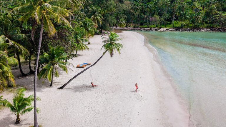 Ein Paar ist an einem Sandstrand umgeben von Palmen und Natur. Die Frau sitzt in einer Schaukel. Der Mann geht Richtung Meer.
