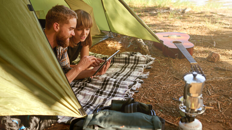 Ein Paar ruht sich im Campingzelt aus, nutzt ein Tablet zur Kommunikation und für Online-Streaming und genießt die gemeinsame Zeit.