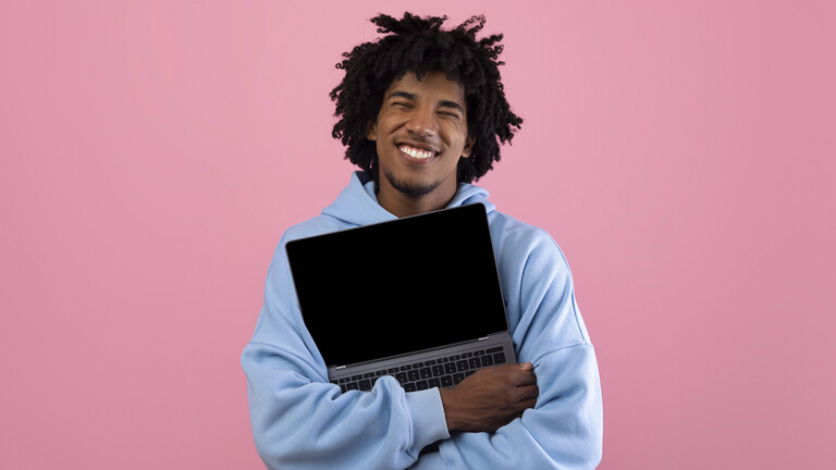 Foto eines jungen Mannes in blauem Pullover vor einer pinken Wand, der ein aufgeklapptes Laptop umarmt