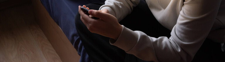 Ein junger Mann sitzt auf seinem Bett und schaut in sein Smartphone