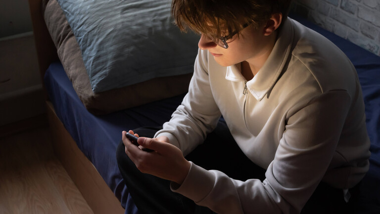 Ein junger Mann sitzt auf seinem Bett und schaut in sein Smartphone