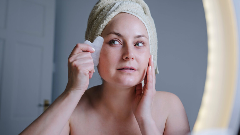 Eine Frau sieht sich selbst in einem Spiegel und massiert ihr Gesicht mit einem Gua Sha, eine Art flacher Stein.