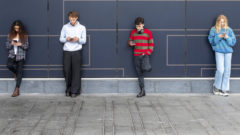 Vier junge Erwachsene stehen angelehnt an eine Wand und schauen jeweils auf ihre Handys.