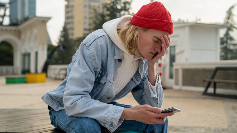Ein junger Typ schaut auf sein Smartphone und hält sich die hand vors Gesicht.