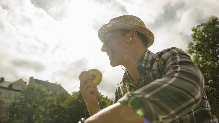 Ein Mann sitzt in der Sonne, trägt einen Strohhut und hält einen Apfel in der Hand (Symbolfoto)