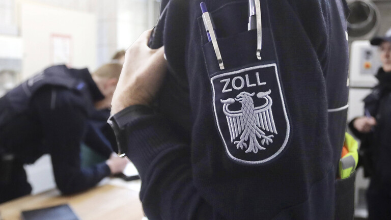 Nahaufnahme des Oberarms eines Zollbeamten mit einem Zoll-Aufnäher auf der Uniform, im Hintegrund sind unscharf Zoll-Beamte bei der Arbeit zu erkennen.