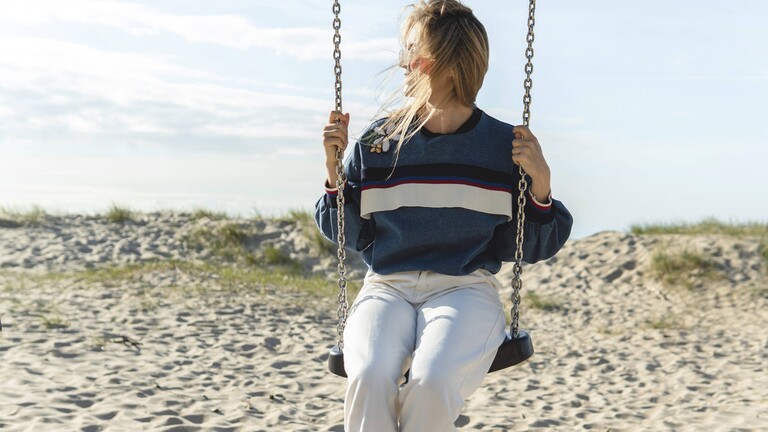 Eine Frau sitzt auf einer Schaukel und schaut zur Seite. Die Schaukel steht an einem Strand. Es scheint die Sonne.