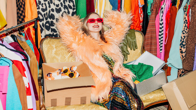 Eine Frau lehnt sich auf einem Sessel zurück. Um sie herum stehen einige Kleiderstangen, die vollgepackt mit Kleidung sind. Die Frau lächelt und trägt eine rote Sonnenbrille.