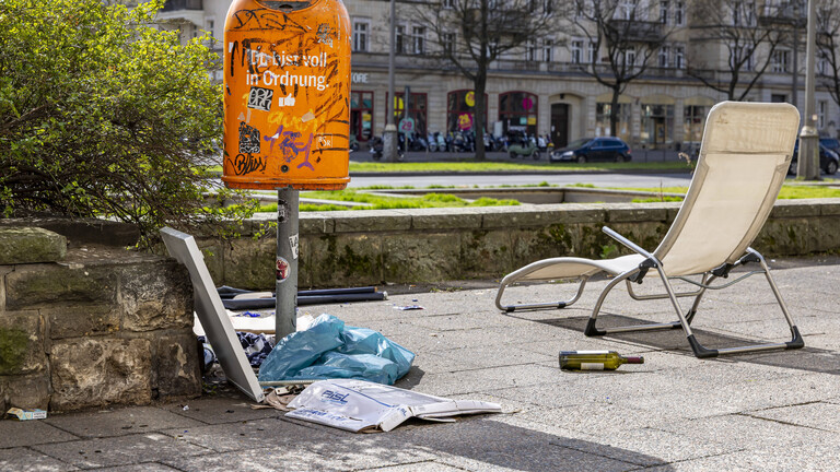 Neben einem Mülleimer auf einer Straße liegt Müll wie Plastiktüten und Sperrmüll wie eine alte Sonnenliege.