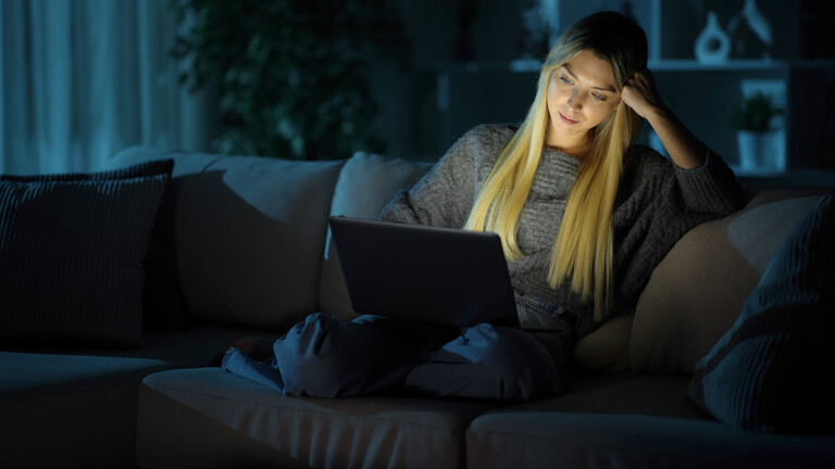 Eine Frau sitzt im Dunkeln auf einer Couch. Sie hat einen Laptop auf ihrem Schoß. Durch die Helligkeit des Bildschirms ist ihr Gesicht auch hell.