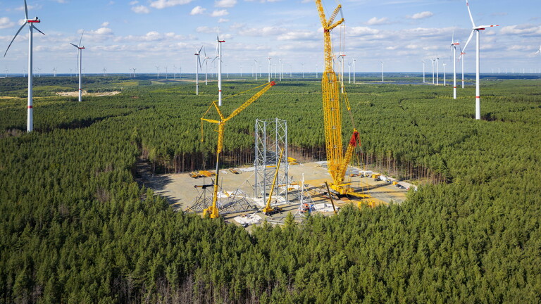 Das Bild zeigt eine Baustelle in einem Wald im brandenburgischen Schipkau. Umgeben von Windrädern wird dort das höchste Windrad der Welt gebaut. Dafür wird eine Gitterkonstruktion gebaut. Um die stehen Kräne.