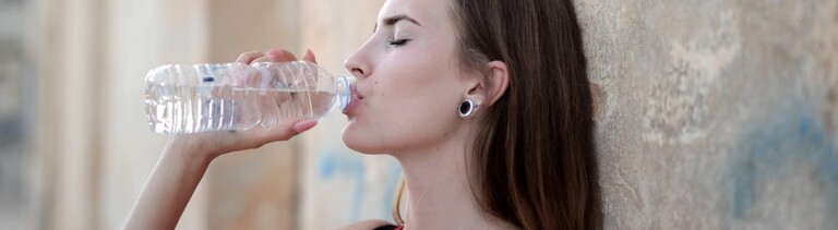 Eine Frau lehnt an einer Mauer und trinkt aus einer Waserflasche.