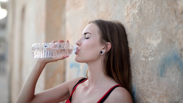 Eine Frau lehnt an einer Mauer und trinkt aus einer Waserflasche.