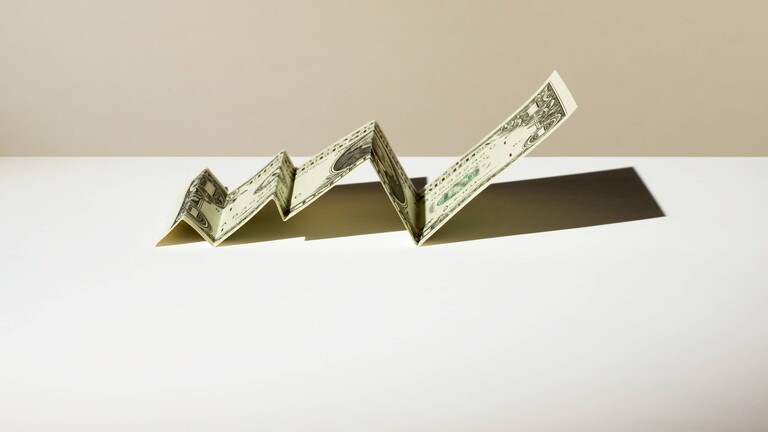 Ein gefalteter Dollar-Schein liegt auf einer Oberfläche. Der Dollar-Schein erinnert durch das Falten an einen Graphen, der ansteigt.