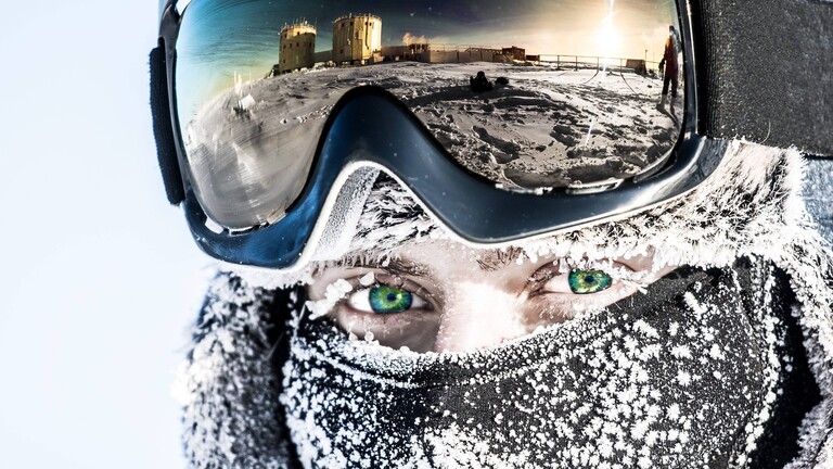 Eine Person in der Antarktis mit entsprechender Schutzausrüstung: einer Art Ski-Brille und einer Gesichtsabdeckung gegen die Kälte. In der Brille spiegelt sich die Concordia-Station.