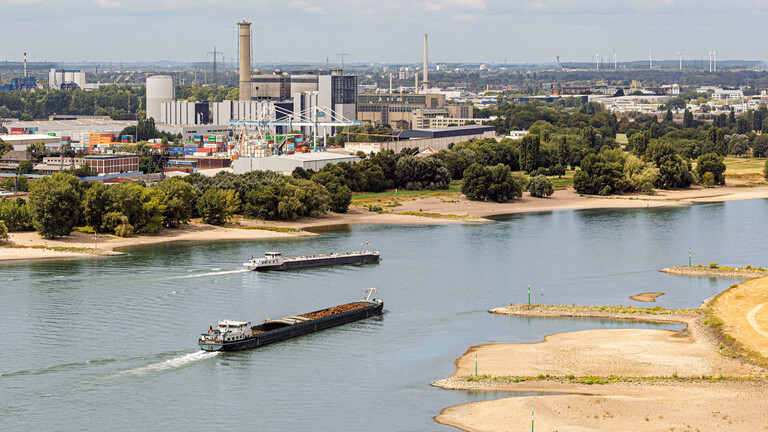 Zwei nicht voll beladende Transport-Binnenschiffe fahren im Juli 2026 durch das Flussbett des Rheins bei Niedrigwasser vor Düsseldorf, im Hintergrund das Heizkraftwerk Lausward.