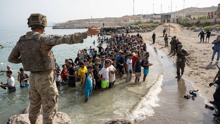 Geflüchtete stehen am Strand von Ceuta. Sie sind über das Meer von der marokkanischen Küste zu der spanischen Enklave geschwommen. Ein Soldat zeigt ihnen den Weg.