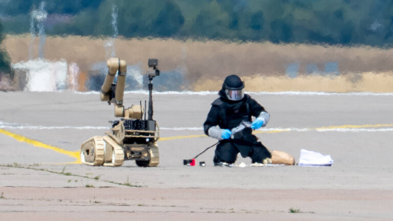 Ein Kriminalbeamter entschärft die Drohne am Flughafen Leipzig/Halle. Neben ihm steht ein Sprengstoffroboter. Der Beamte trägt Schutzkleidung und einen Helm mit Visier.