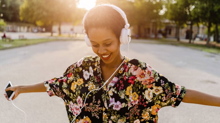 Foto einer jungen Frau, die mit geschlossenen Augen und lächelnd auf einem Kopfhörer Musik hört und die Arme ausgebreitet hat, in der rechten Hand ein Smartphone