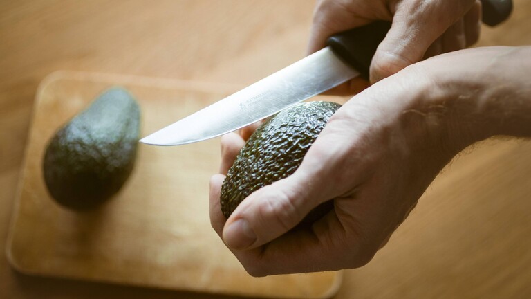 Nahaufnahme von Männerhänden, die mit einem großen Küchenmesser eine Avocado aufschneiden, das Messer in der rechten, die Avocado in der linken Hand