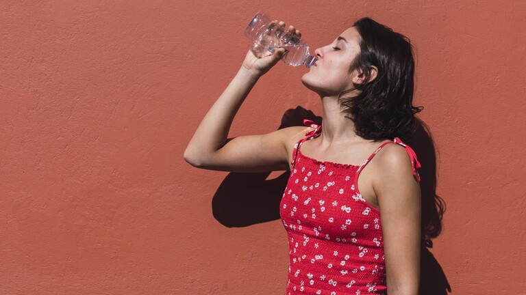 Eine junge Frau trinkt vor einer roten Wand im Sommer aus einer Wasserflasche.