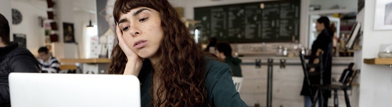 Eine Frau sitzt in einem Café vor einem aufgeklappten Laptop. Sie hat ihren Kopf auf einer Hand abgestützt und schaut gelangweilt auf den Bildschirm.