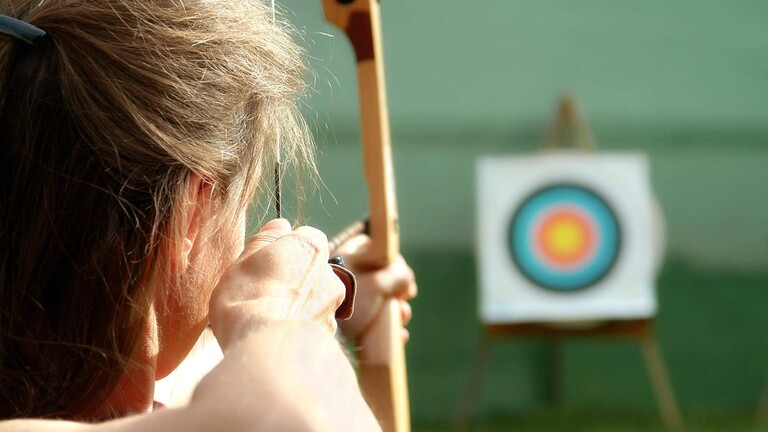 Eine Frau zielt mit Pfeil und Bogen auf eine bunte Zielscheibe.