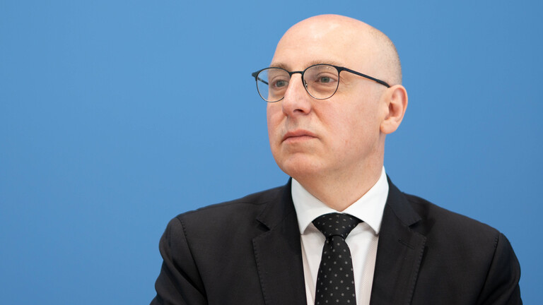 Ein Mann im Anzug mit Brille sitzt vor einer blauen Wand und guckt zum linken Bildrand